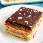 A rectangular dessert bar with layers of shortbread, caramel, and chocolate, topped with flaky sea salt, served on a white plate.