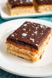A rectangular dessert bar with layers of shortbread, caramel, and chocolate, topped with flaky sea salt, served on a white plate.