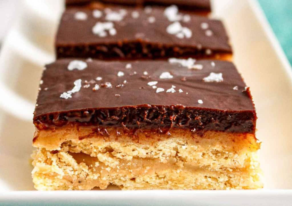 Close-up of chocolate caramel shortbread bars topped with flaky sea salt, arranged in a row on a white plate.