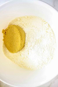 A white bowl containing a mound of flour and a scoop of brown sugar placed next to each other.