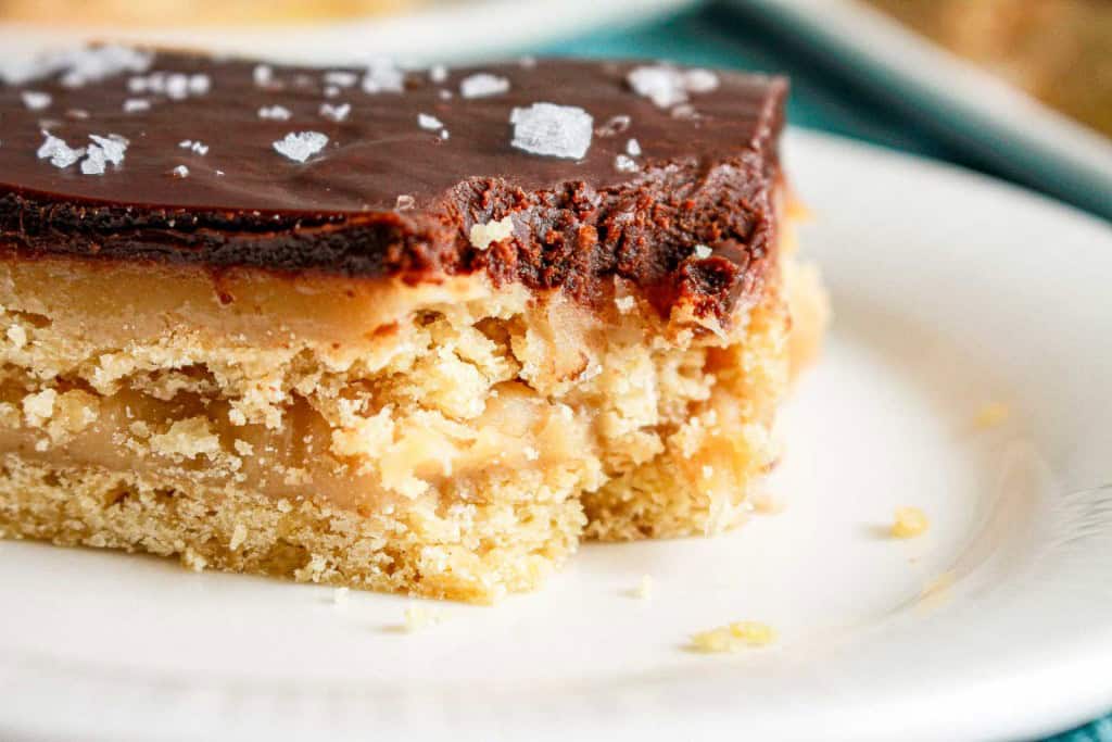 A close-up of a dessert bar with a crumbly crust, caramel layer, chocolate topping, and sprinkled sea salt on a white plate.