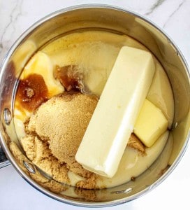 A saucepan containing a stick of butter, brown sugar, sweetened condensed milk, and vanilla extract on a marble countertop.