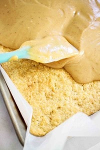 A blue spatula spreads a thick, light brown frosting over a baked crumbly crust in a parchment-lined baking pan.