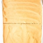 A large rectangular dessert with a smooth light brown frosting, sliced once across the middle, sits on white parchment paper.