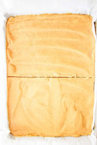 A large rectangular dessert with a smooth light brown frosting, sliced once across the middle, sits on white parchment paper.