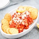 Oval dish with a mixture of chopped onions, tomato-based sauce, and a lemon wedge in the center, surrounded by round crackers on a gray surface.