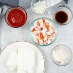 Five bowls contain ketchup, chopped onions, Worcestershire sauce, chopped imitation crab, and mayonnaise beside a plate with two blocks of cream cheese on a gray surface.