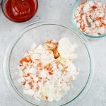 A glass bowl with chopped imitation crab, mayonnaise, and diced onions, with a side bowl of ketchup and another bowl of chopped crab on a gray surface.