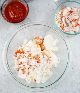 A glass bowl with chopped imitation crab, mayonnaise, and diced onions, with a side bowl of ketchup and another bowl of chopped crab on a gray surface.