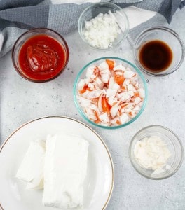 Five bowls contain ketchup, chopped onions, Worcestershire sauce, chopped imitation crab, and mayonnaise beside a plate with two blocks of cream cheese on a gray surface.