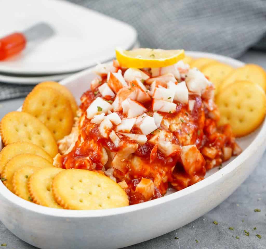 A white dish with a cheese ball topped with chopped onions, cocktail sauce, and a lemon slice, surrounded by round crackers.