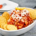 A white dish with a cheese ball topped with chopped onions, cocktail sauce, and a lemon slice, surrounded by round crackers.