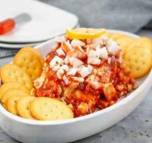 A white dish with a cheese ball topped with chopped onions, cocktail sauce, and a lemon slice, surrounded by round crackers.