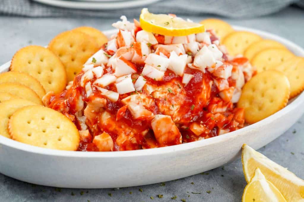 A white dish filled with a crab dip topped with cocktail sauce and diced crab meat, garnished with a lemon slice, surrounded by round crackers.