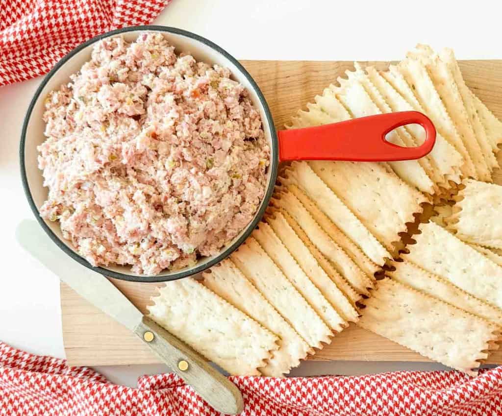 A bowl of ham salad with a red handle, surrounded by rectangular crackers on a wooden board, with a spreader knife and red checkered cloth nearby.
