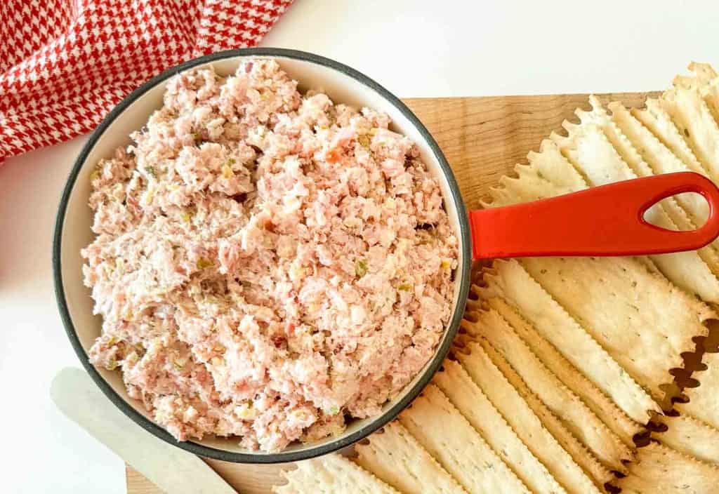 A bowl of ham salad with a red handle beside rows of rectangular crackers on a wooden board, with a red and white cloth in the background.