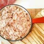 A bowl of ham salad with a red handle beside rows of rectangular crackers on a wooden board, with a red and white cloth in the background.