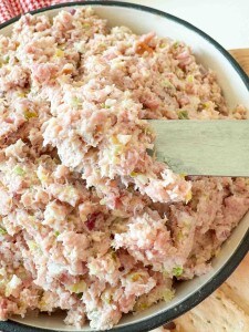 A bowl filled with a mixture of finely chopped ham, pickles, and mayonnaise, being stirred with a wooden spatula.