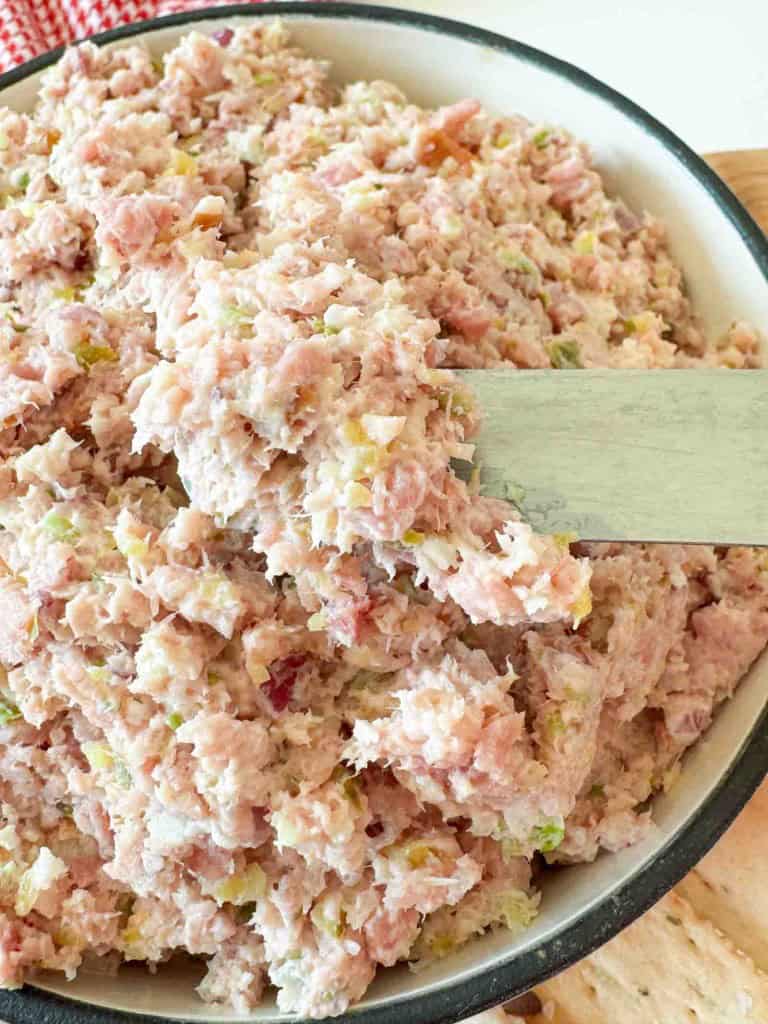 A bowl filled with a mixture of finely chopped ham, pickles, and mayonnaise, being stirred with a wooden spatula.