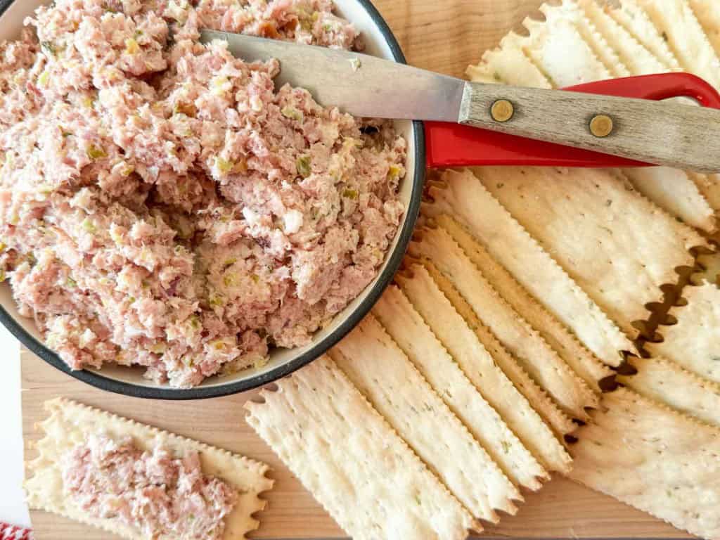 A bowl of ham salad spread with a knife next to a stack of rectangular crackers on a wooden surface.
