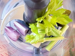 Chopped red onion, celery stalks, and celery leaves in a food processor with a metal blade attachment.