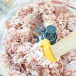 Ground meat mixture with chopped vegetables and white ingredients being mixed in a food processor with a yellow spatula.