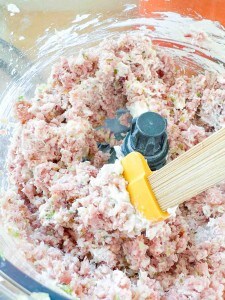 Ground meat mixture with chopped vegetables and white ingredients being mixed in a food processor with a yellow spatula.