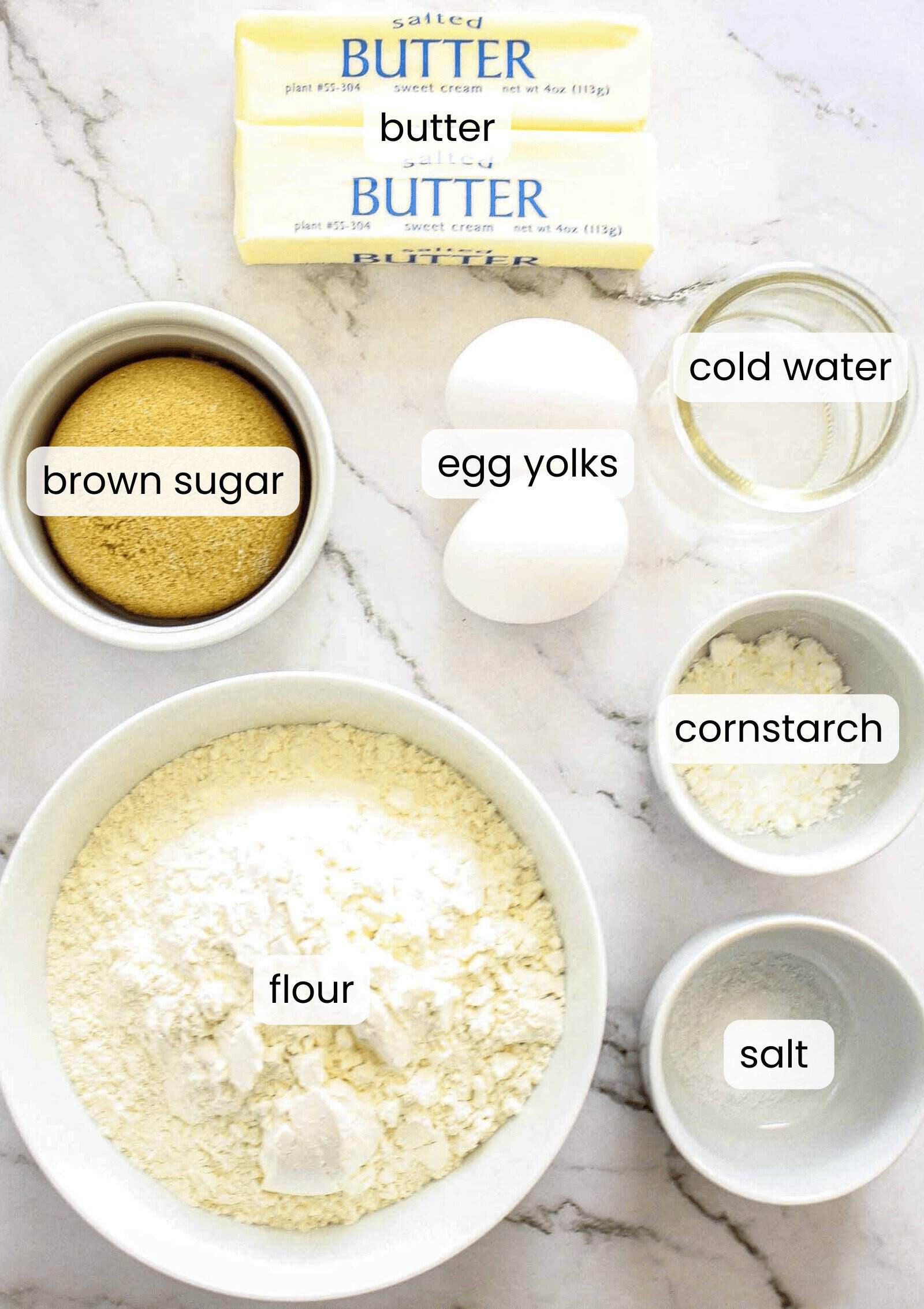 Top-down view of baking ingredients for Caramel Chocolate Bars on a marble surface, featuring labeled bowls of brown sugar, flour, cornstarch, salt, two eggs, cold water, and two sticks of salted butter.