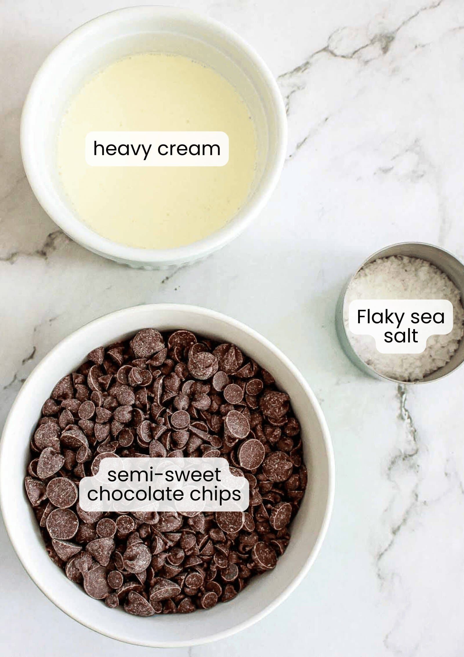 A bowl of heavy cream, semi-sweet chocolate chips, and a small container of flaky sea salt are ready on a marble surface&mdash;perfect for making decadent Caramel Chocolate Bars.