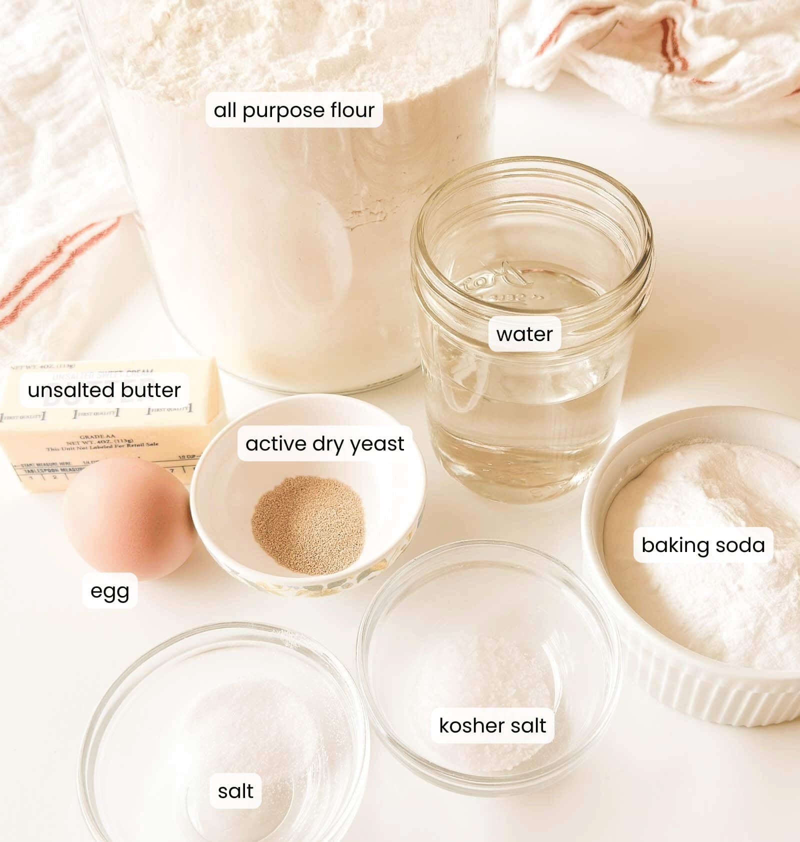 Ingredients for baking displayed on a white surface: all purpose flour, water, unsalted butter, egg, active dry yeast, baking soda, kosher salt, and salt, each labeled.