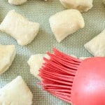 A red silicone brush applies egg wash to raw dough pieces on a baking sheet lined with a textured mat.