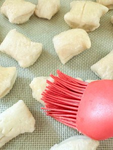 A red silicone brush applies egg wash to raw dough pieces on a baking sheet lined with a textured mat.