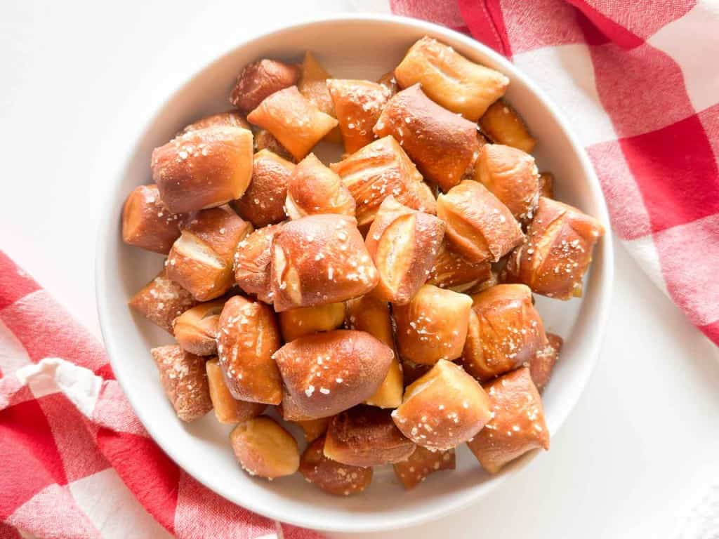 A white bowl filled with salted soft pretzel bites sits on a surface next to red and white checkered cloths.