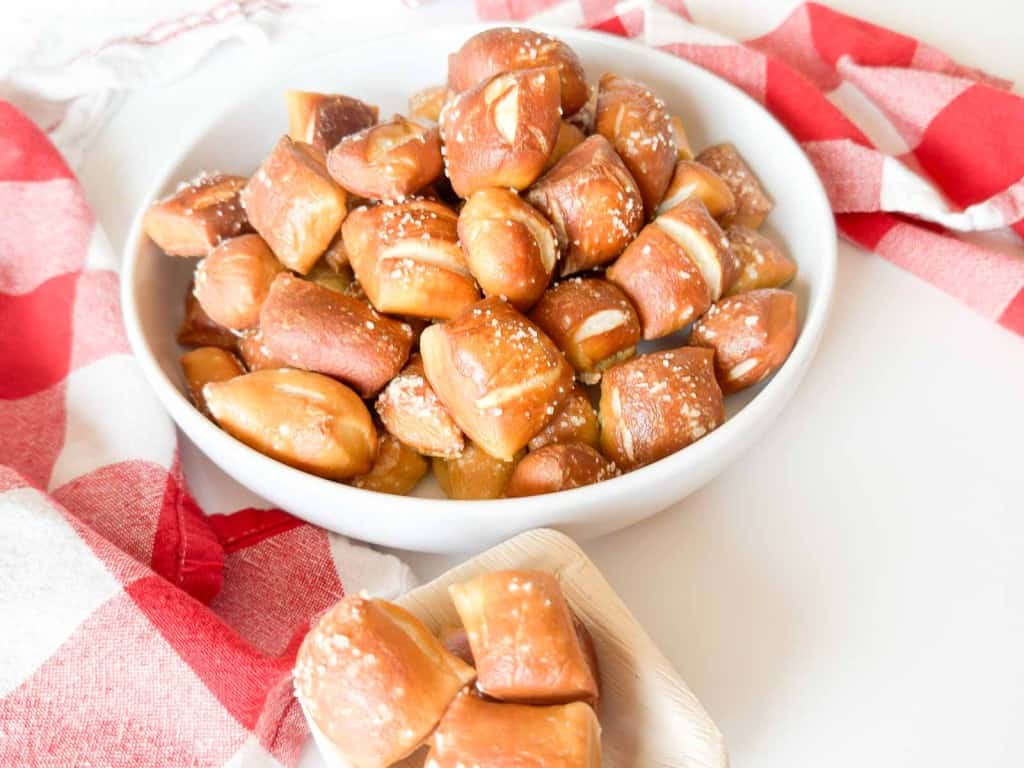 A white bowl filled with soft pretzel bites topped with coarse salt, placed on a white surface next to a red and white checkered cloth.