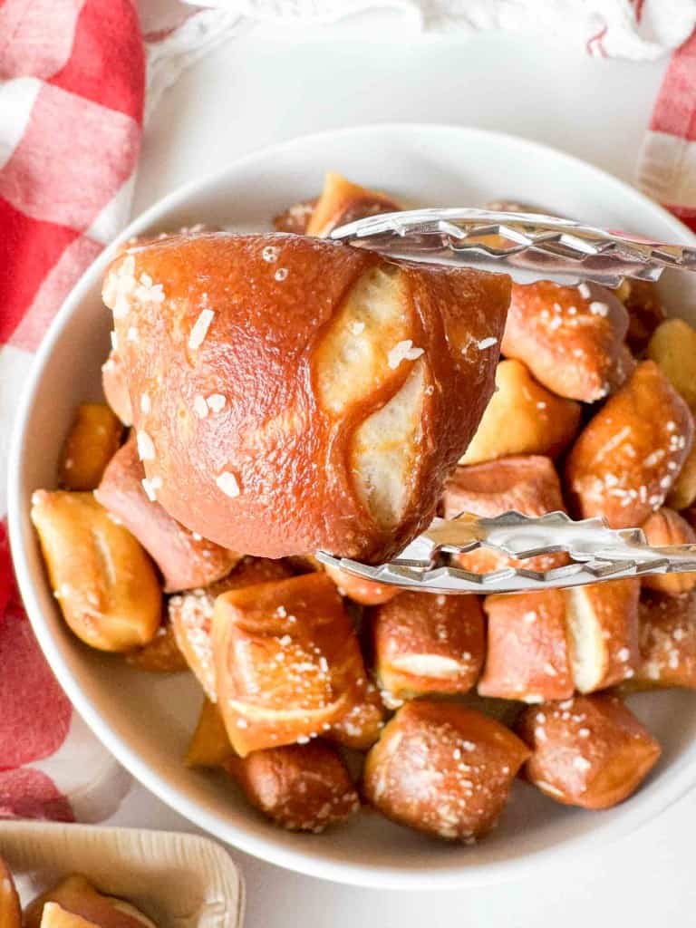 A pair of metal tongs holds a salted pretzel bite above a white bowl filled with more pretzel bites, next to a red and white checkered cloth.
