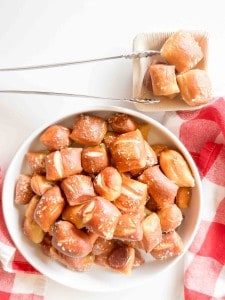 A bowl of soft pretzel bites sprinkled with coarse salt, with a small serving of pretzel bites on a wooden plate and metal tongs beside it.