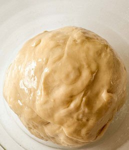 A ball of unbaked dough sits in a white bowl, lightly coated with oil.