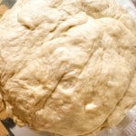 Ball of risen dough on a clear glass plate, showing a smooth and slightly textured surface.