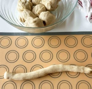A rolled log of dough on a baking mat with circle guides, with a glass bowl of dough balls and a towel in the background.