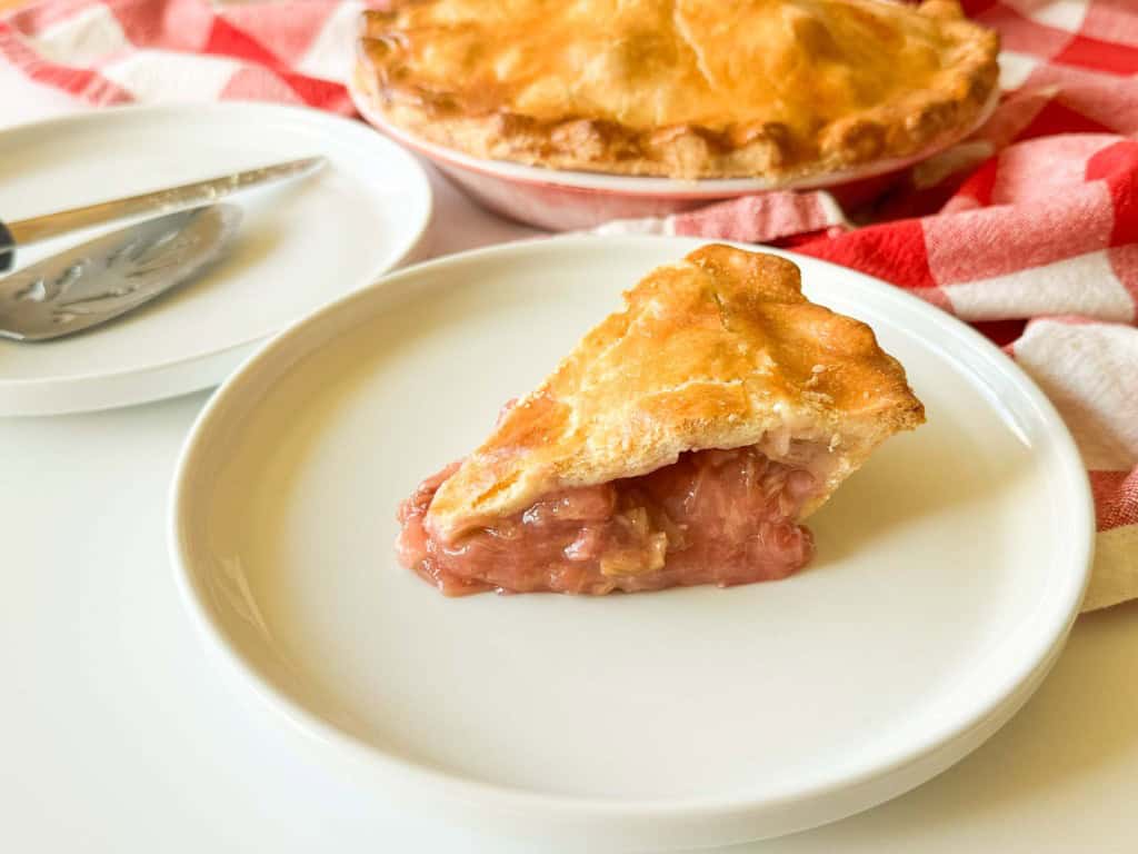 A slice of classic rhubarb pie sits on a white plate, with a serving utensil resting on a nearby empty plate and the whole pie in the background atop a red and white checkered cloth.