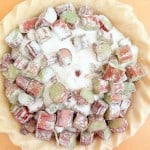 Unbaked classic rhubarb pie crust filled with chopped rhubarb pieces, topped with a layer of granulated sugar and flour, rests on a wooden surface.