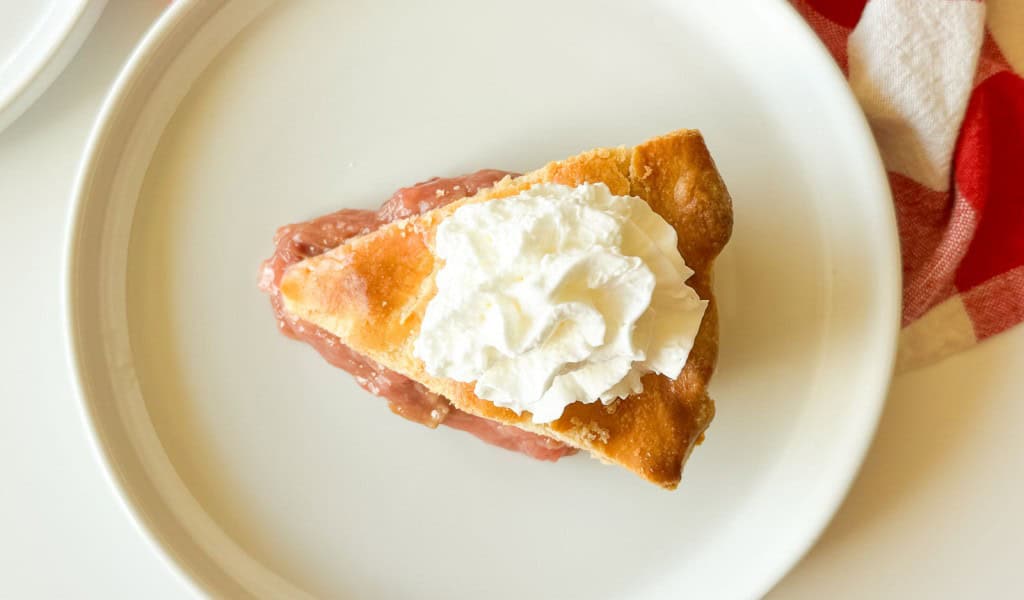 A slice of classic rhubarb pie with a golden crust and pink filling is topped with whipped cream on a white plate.