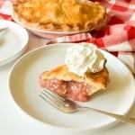 A slice of classic rhubarb pie with a dollop of whipped cream on a white plate, fork beside it, the whole pie and a red-checkered cloth visible in the background.