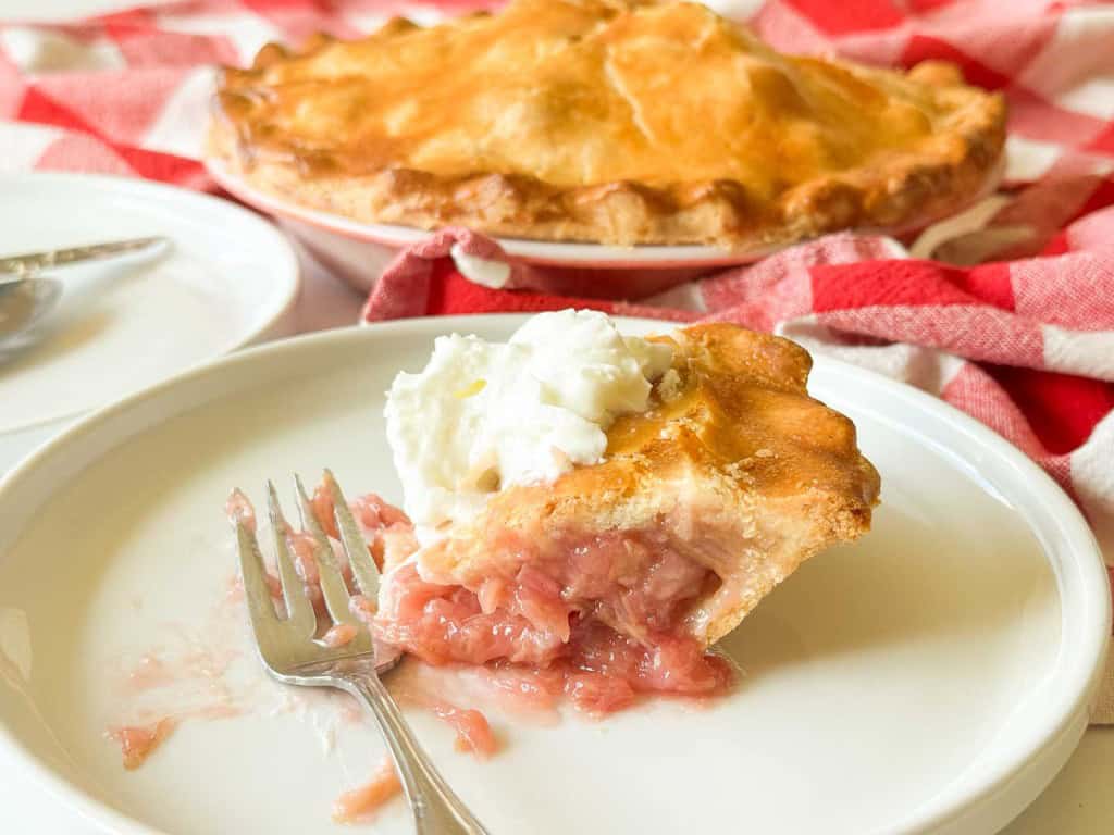 A slice of classic rhubarb pie with a dollop of whipped cream sits on a plate, accompanied by a fork and the rest of the pie in the background.