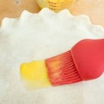 A red silicone brush applies an egg wash to a classic rhubarb pie crust; a Pyrex measuring cup with yellow liquid is in the background.