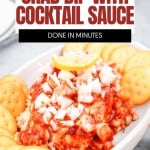 A dish of Crab Dip with Cocktail Sauce, topped with chopped onions and garnished with a lemon slice, is surrounded by round crackers on a white tray.