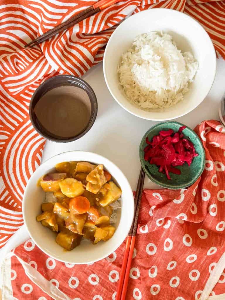 A bowl of chicken curry with rice, pickled ginger, and a beverage, served with chopsticks and festive napkins.