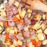 Raw chicken pieces, diced potato, sliced carrots, and onion cooking in a stainless steel pan, stirred with a wooden spatula.