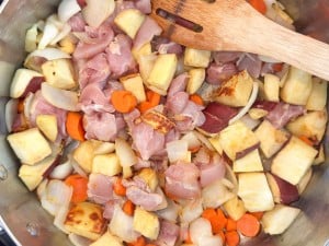 Raw chicken pieces, diced potato, sliced carrots, and onion cooking in a stainless steel pan, stirred with a wooden spatula.