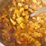 A ladle scoops chicken curry with potatoes, carrots, and onions from a metal pot.